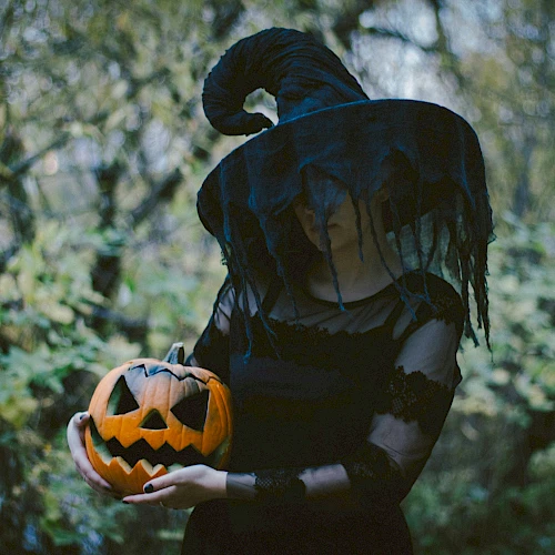 A person in a witch costume holds a carved pumpkin outdoors, surrounded by trees and foliage.