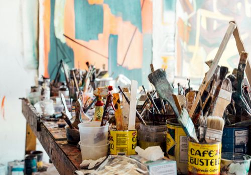 The image shows a cluttered art studio with paintbrushes, various art supplies, and a colorful abstract painting in the background.