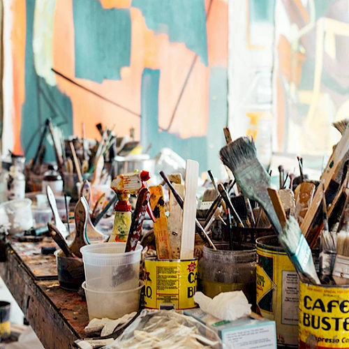 The image shows a cluttered art studio with paintbrushes, various art supplies, and a colorful abstract painting in the background.
