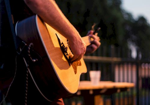 A person is playing an acoustic guitar outdoors, with a blurred background of trees and a cup on a table.