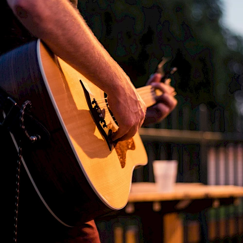A person is playing an acoustic guitar outdoors, with a blurred background of trees and a cup on a table.