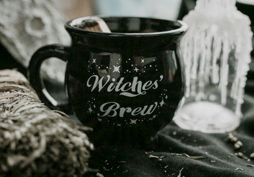 A black mug labeled "Witches' Brew" with stars, surrounded by a sage bundle and a candle on a dark fabric.