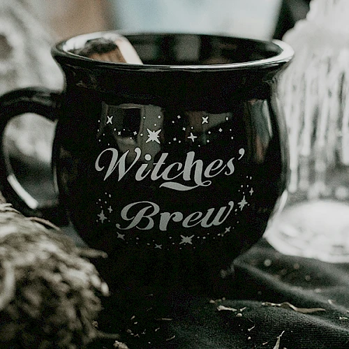 A black mug labeled "Witches' Brew" with stars, surrounded by a sage bundle and a candle on a dark fabric.