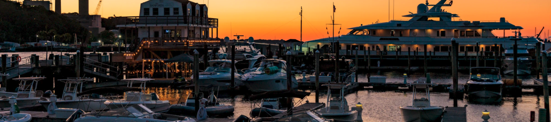 A marina at sunset with boats docked and a large yacht in the background, set against an orange and blue sky.