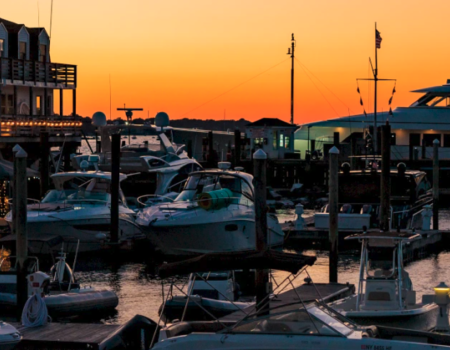A marina at sunset with boats docked and a large yacht in the background, set against an orange and blue sky.