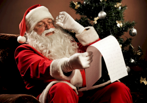 Santa Claus is sitting by a Christmas tree, reading a list, and wearing his traditional red suit and hat.