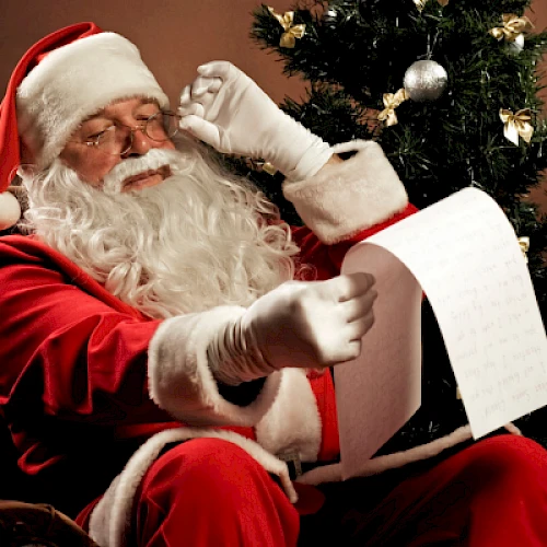 Santa Claus is sitting by a Christmas tree, reading a list, and wearing his traditional red suit and hat.