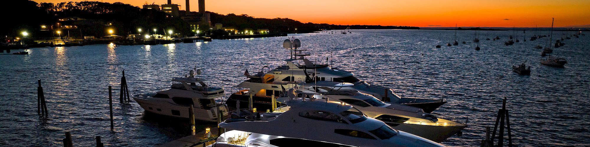 Boats docked at a marina at dusk, calmed water, city lights, orange-to-navy sunset with smokestacks in the distance, serene evening harbor scene.