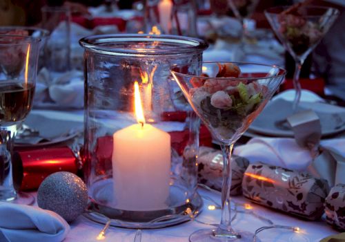 A candlelit table with wine glasses, a martini glass, wrapped gifts, and a soft glow from a centerpiece candle, set for a festive dinner.