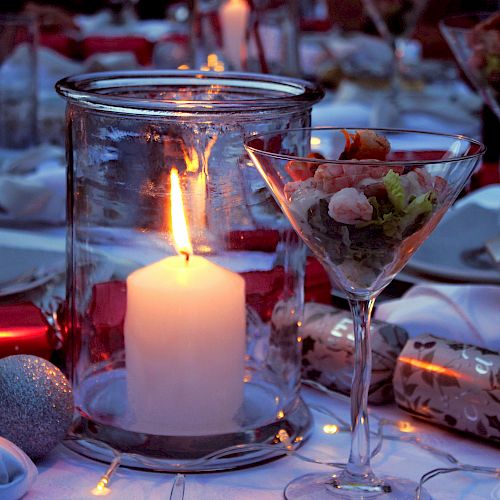 A candlelit table with wine glasses, a martini glass, wrapped gifts, and a soft glow from a centerpiece candle, set for a festive dinner.