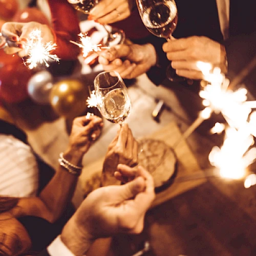 People celebrate with sparks, drinks, and cheers, toasts raised high as festive sparks fly and confetti sparkles illuminate the scene.