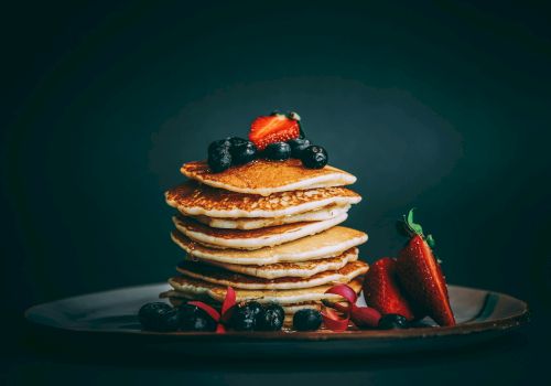 A stack of golden pancakes topped with berries sits on a dark plate, with fresh strawberries and blueberries arranged at the base.