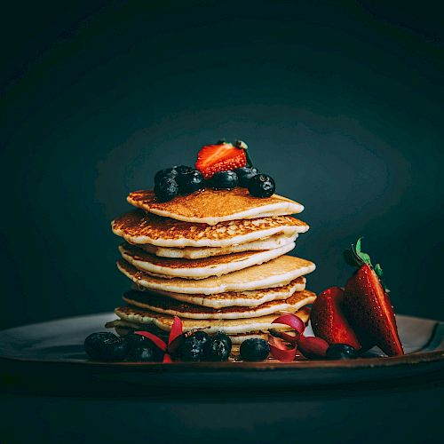 A stack of golden pancakes topped with berries sits on a dark plate, with fresh strawberries and blueberries arranged at the base.