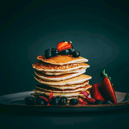 A stack of golden pancakes topped with berries sits on a dark plate, with fresh strawberries and blueberries arranged at the base.