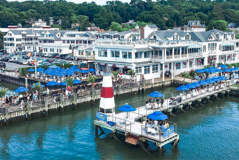 A waterfront boardwalk with white seaside buildings, a docked pier, blue umbrellas, and a small water taxi platform by the harbor.