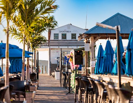 A sunny boardwalk cafe scene with blue umbrellas, palm trees, and a white building in the distance; chairs line the wooden deck, inviting vibes.