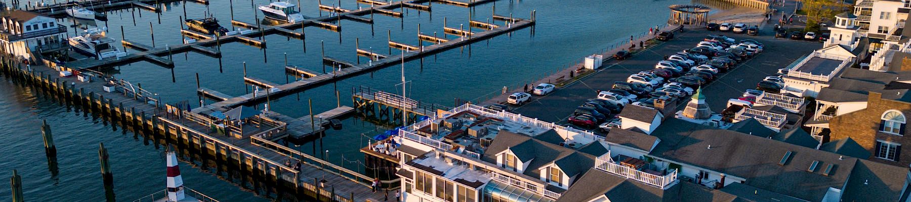 A coastal marina with numerous boats, a long pier, cluster of white houses, and calm blue water reflecting a sunny sky.