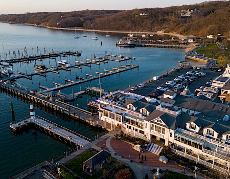 A coastal marina with numerous boats, a long pier, cluster of white houses, and calm blue water reflecting a sunny sky.