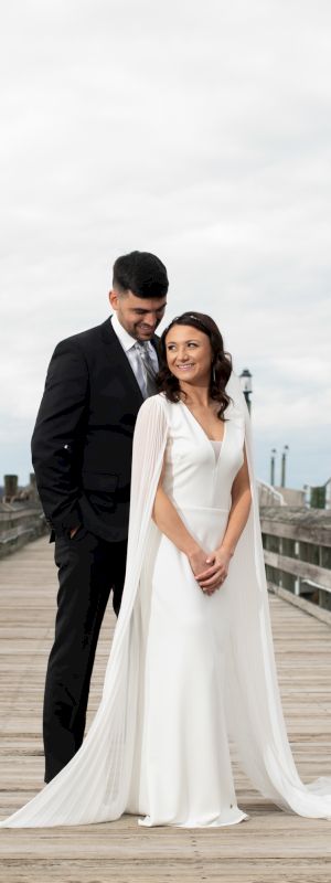 A newlywed couple stands on a wooden pier in elegant attire, the man in a black suit and the woman in a flowing white gown, smiling together.
