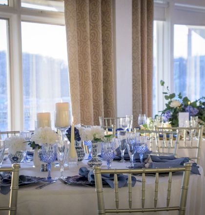 A formal banquet hall with round tables draped in white, blue accents, elegant chairs, floral centerpieces, large windows, and soft natural light.