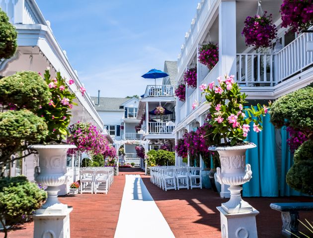 A sunny, narrow Mediterranean street with white buildings, colorful doors, and potted plants along a tiled path, evoking a cheerful coastal vibe.