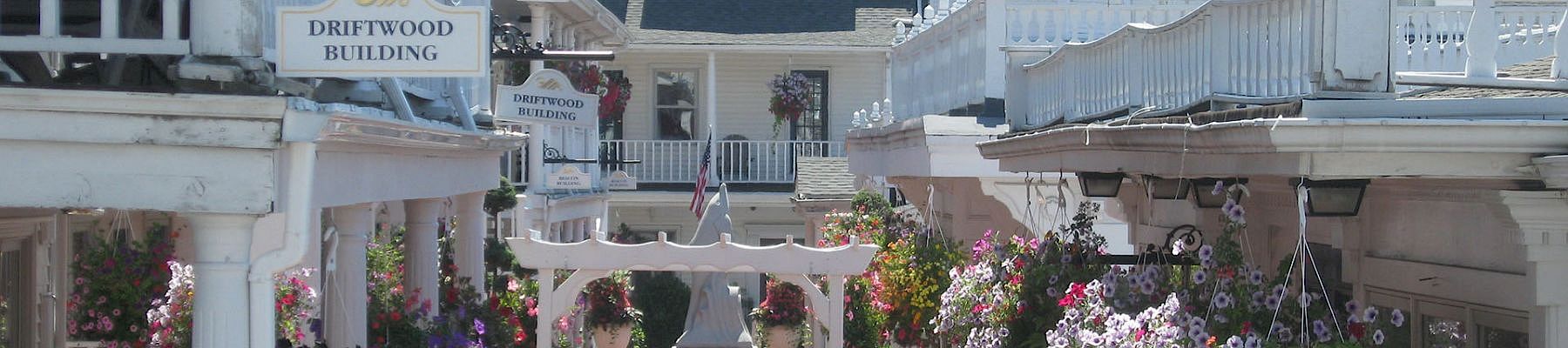 A charming seaside village street with white buildings, flower baskets, and quaint storefronts lining a sunny brick walkway.