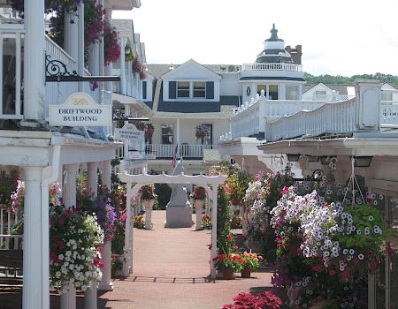 A charming seaside village street with white buildings, flower baskets, and quaint storefronts lining a sunny brick walkway.