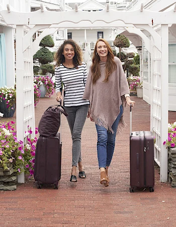 Two smiling women walk through a white archway with rolling suitcases, flowers, and a pleasant resort-like entrance, posing for travel photos.