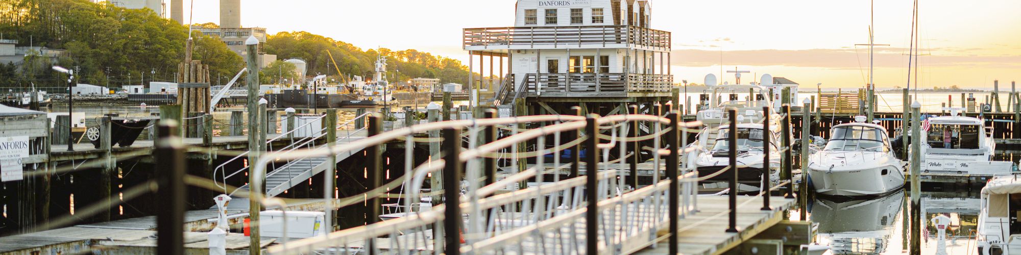 A wooden dock with small boats tied around, calm water, and a white building at the end reflecting a sunset.