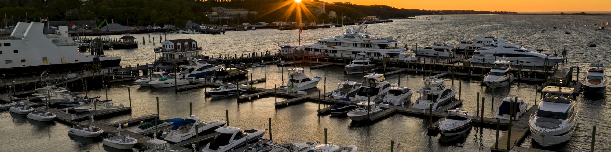 Aerial view of a busy marina at sunset, with numerous boats docked and calm water reflecting the orange sky and silhouettes of distant shore.