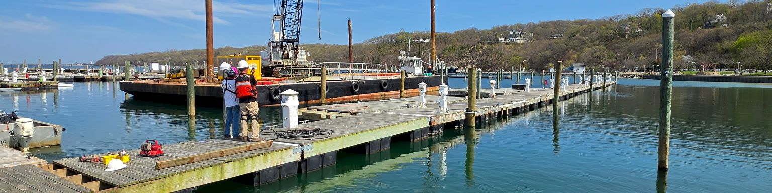 A marina with a small dock, a crane on a floating platform, and calm green-blue water under a clear blue sky.