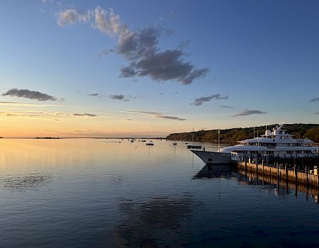 Boats docked by a calm harbor at sunset, reflecting orange and blue skies over still water, a peaceful seaside scene with distant hills.