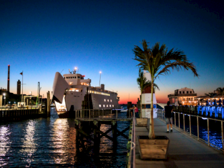 A seaside dock at dusk with a modern ferry, palm tree, calm water, and lit buildings along the pier. End.