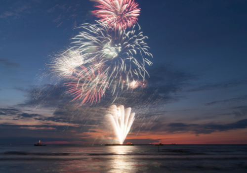 Fireworks burst over a dark, calm sea at dusk, colorful explosions lighting the sky near the horizon, reflecting on the water&rsquo;s surface.
