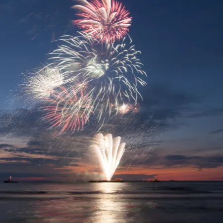 Fireworks burst over a dark, calm sea at dusk, colorful explosions lighting the sky near the horizon, reflecting on the water&rsquo;s surface.