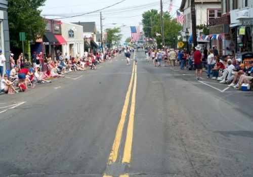 A parade on a small-town street with crowds lining both sides, spectators seated, American flags, and a float approaching in the distance.