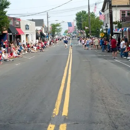 A parade on a small-town street with crowds lining both sides, spectators seated, American flags, and a float approaching in the distance.