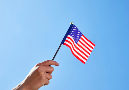 A hand holds a small US flag against a clear blue sky, waving proudly.
