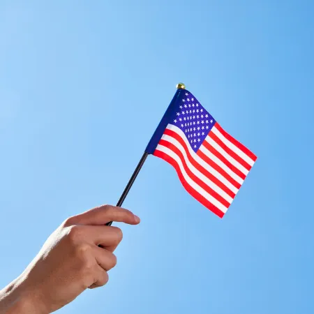 A hand holds a small US flag against a clear blue sky, waving proudly.