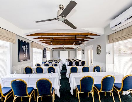 A banquet hall with long tables, blue-and-gold chairs, white tablecloths, ceiling fans, and windows with blinds.