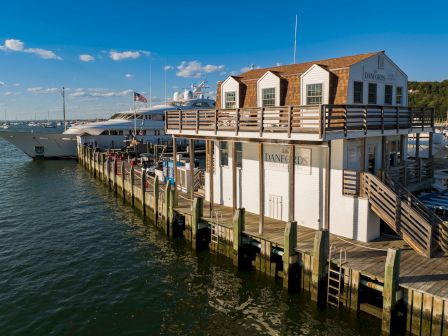 A waterfront dock with the Danfords Hotel & Marina building beside a large ship, set on calm water under a blue sky.