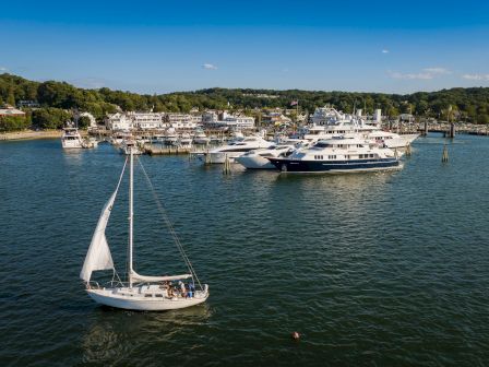 A sailboat and a large yacht are docked along a marina waterfront with houses and lush green hills under a blue sky.