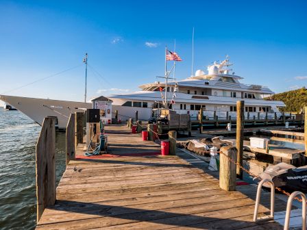 A luxury yacht with an American flag is docked at a wooden pier by the water under a clear blue sky.