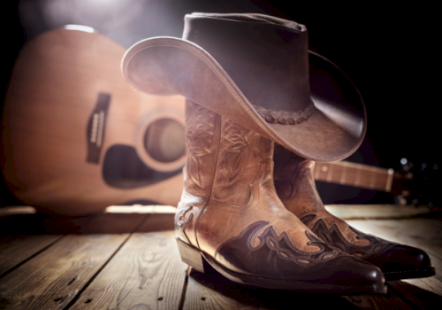 A pair of western cowboy boots with a hat on a wooden floor, with a guitar blurred in the background.