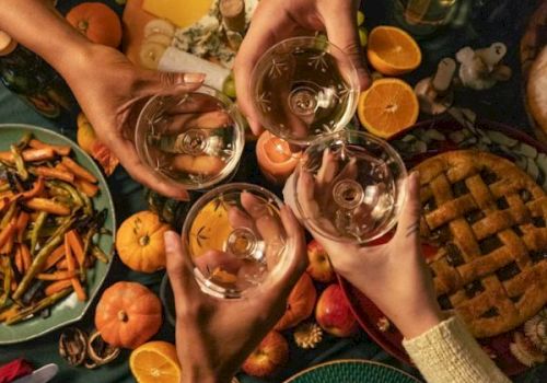 A group of people clink glasses of wine over a table filled with fruits, pumpkins, and snacks, celebrating together.