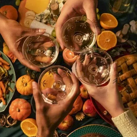 A group of people clink glasses of wine over a table filled with fruits, pumpkins, and snacks, celebrating together.