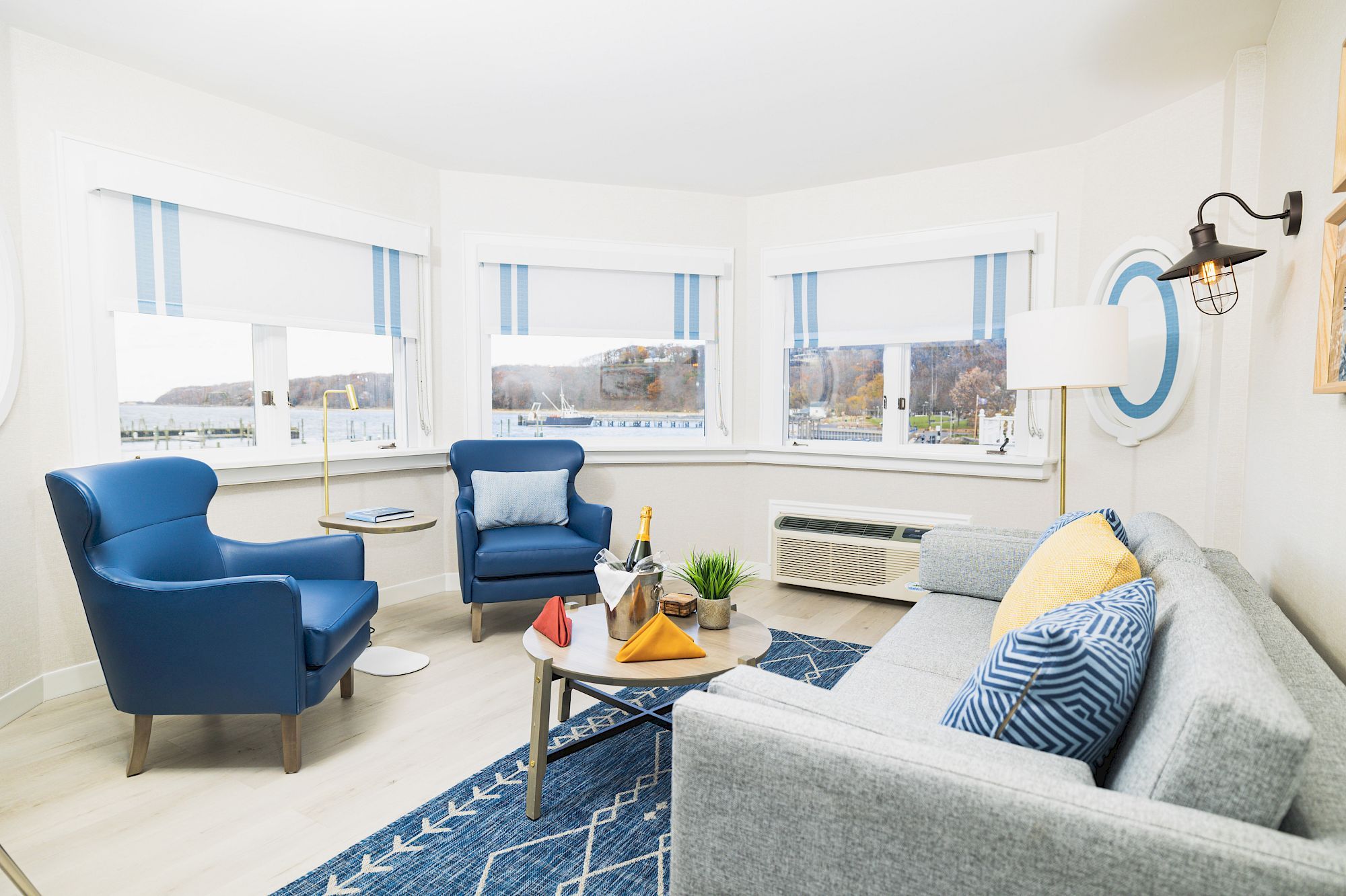 A bright living room with blue chairs, a grey sofa, a blue rug, and a coffee table with decor. It features large windows with striped blinds.