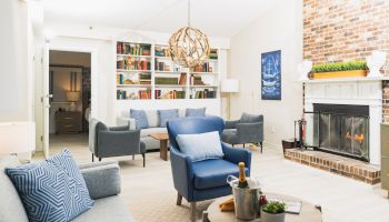 A cozy living room with a fireplace, blue armchairs, a bookshelf, and a chandelier. Bottled champagne with two glasses on a coffee table.