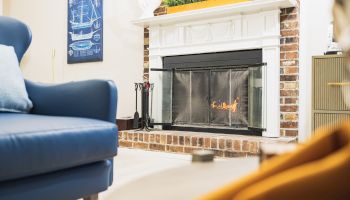 A cozy living room with a fireplace, a blue armchair, a wall art piece, and a plant on the mantle. There's also a hint of an orange item.