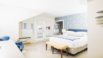 A modern, bright hotel room with a white and blue color scheme, featuring a large bed, a bench, chairs, a desk, and large windows with blinds.